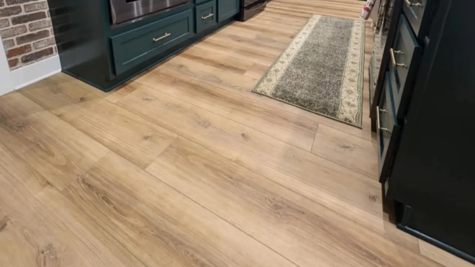 Light oak-look WPC luxury vinyl plank flooring installed through a finished kitchen with deep green cabinets and a brick backsplash, a Miller's Wholesale Flooring install