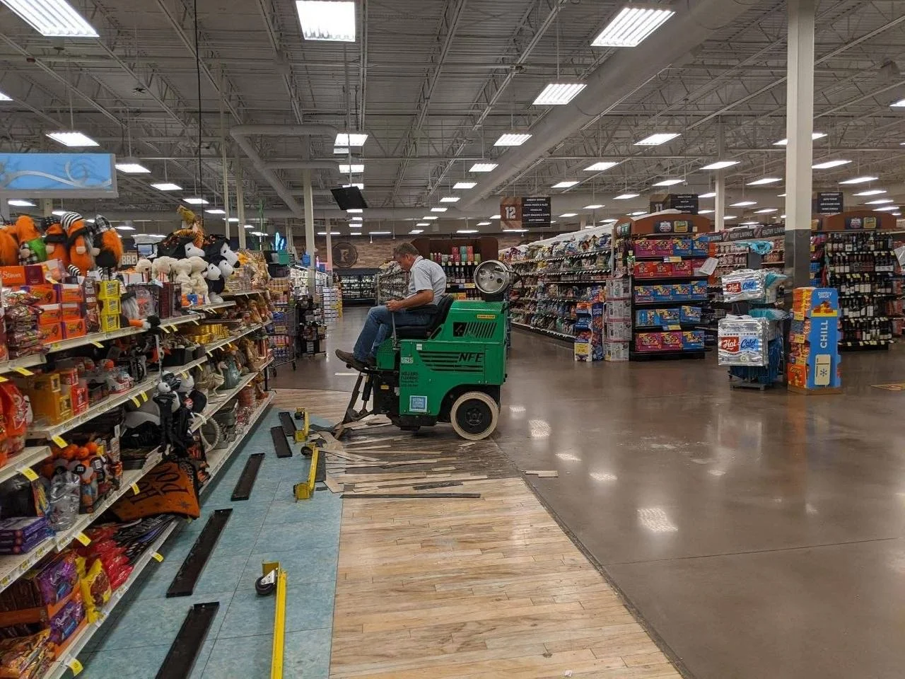 Miller’s operator using a green ride-on floor scraper to lift flooring in a grocery store aisle, with removed strips and the exposed surface visible.